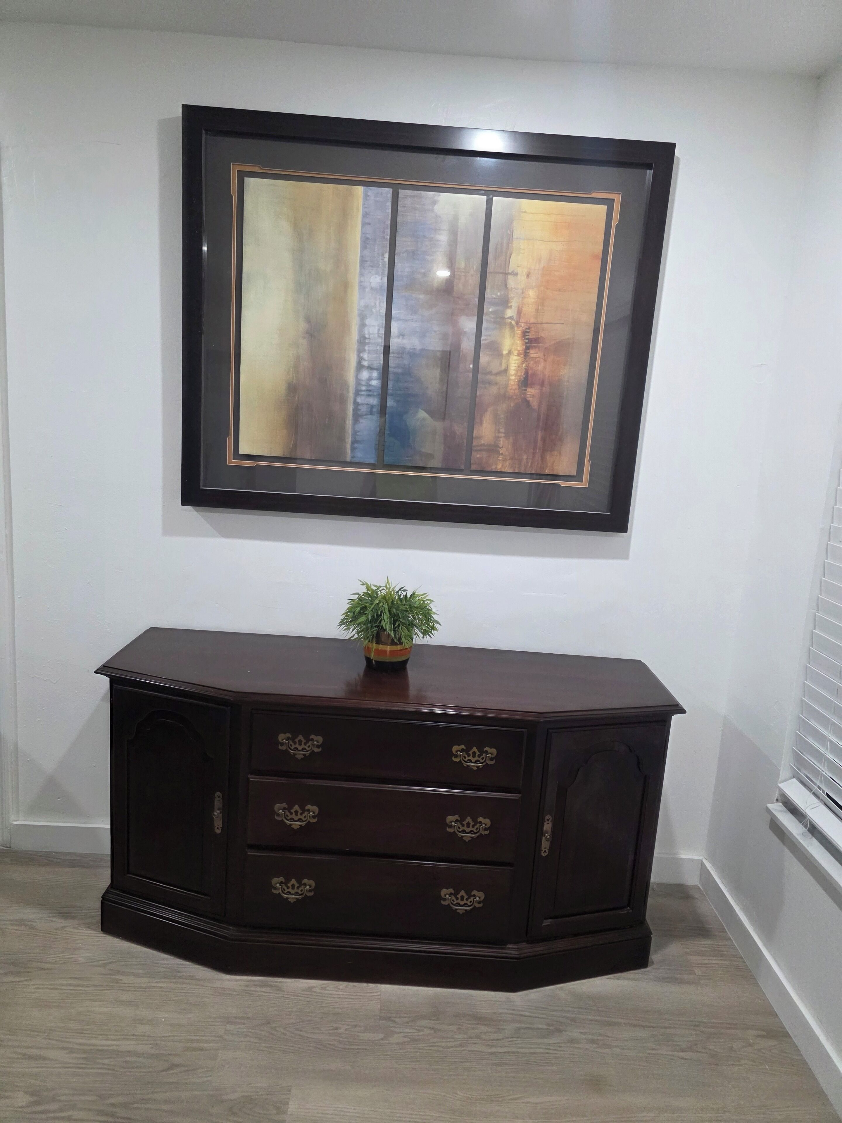 A dark wooden cabinet with ornate handles, a framed abstract painting above it, and a small potted plant on top.