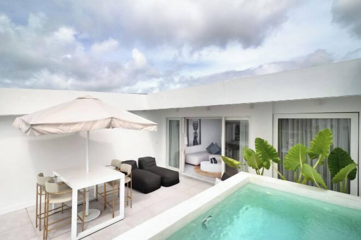 Un patio exterior moderno con piscina, zona de comedor y espacio de descanso con una sombrilla grande.