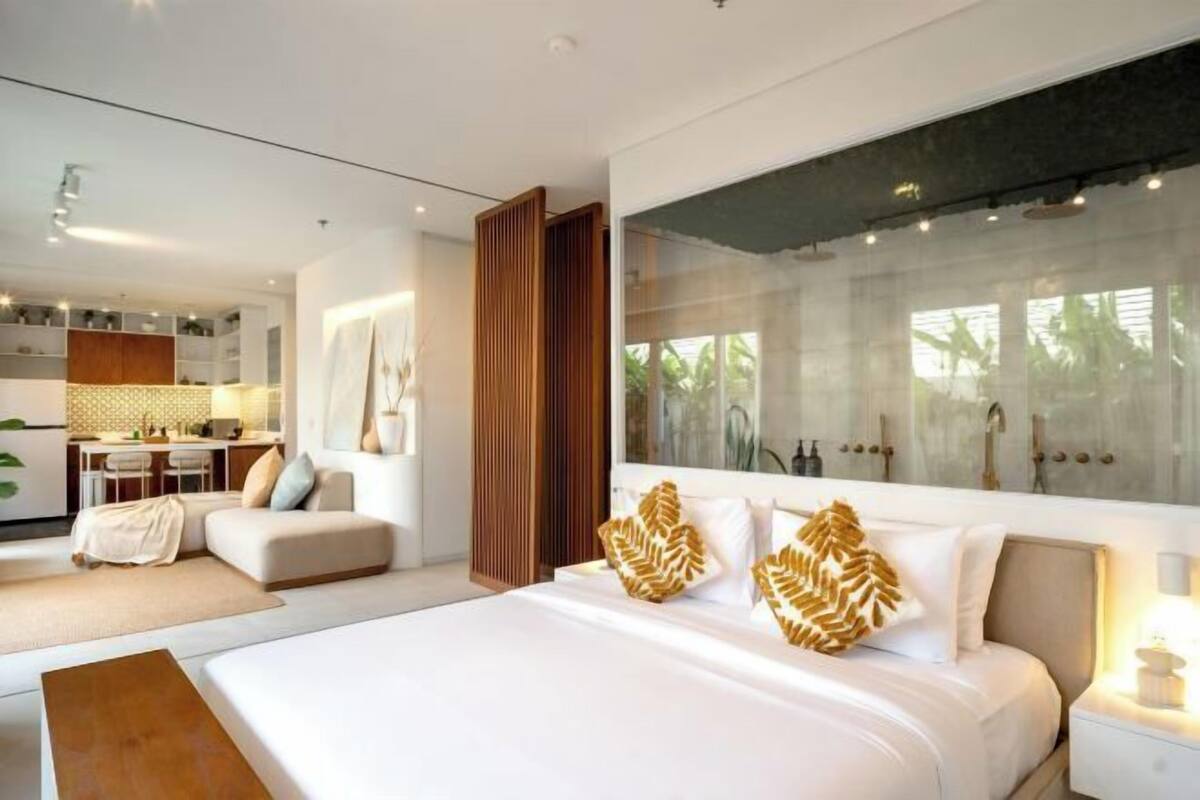 Habitación de hotel moderna con una cama grande, zona de estar con sofá y sillones, kitchenette y un ventanal con vistas a palmeras.