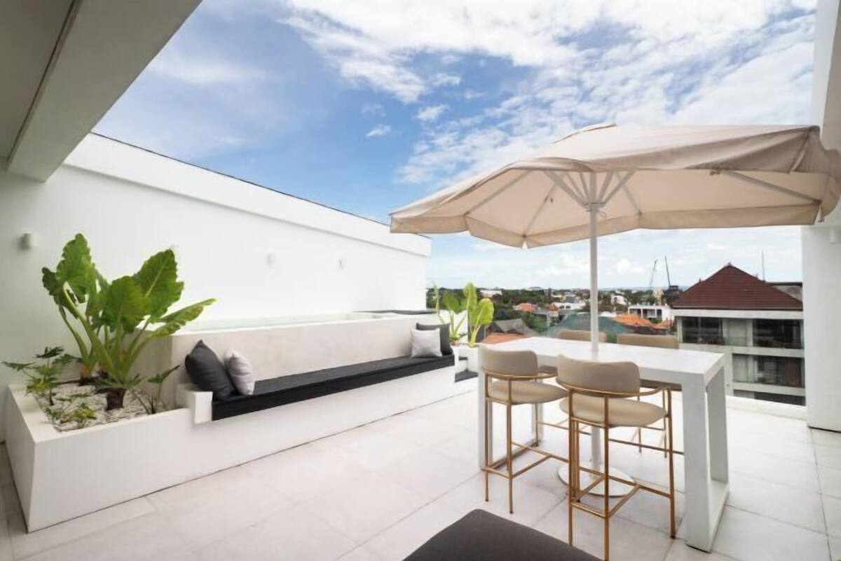 Un área de estar moderna al aire libre con un sofá blanco, una mesita y dos taburetes altos. Un paraguas grande ofrece sombra.