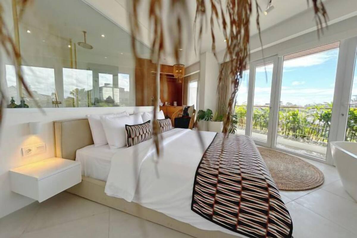 Un dormitorio moderno con una cama grande, mesitas de noche, un banco estampado y vistas al exterior.