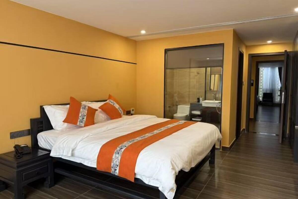A hotel room with a large bed, a bedside table with a phone, a glass door leading to a bathroom, and a wooden floor.