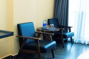 A hotel room with two blue armchairs, a small round table with water bottles and glasses, and a view of the city through a window with curtains. - AN HOTEL (Bac Ninh)