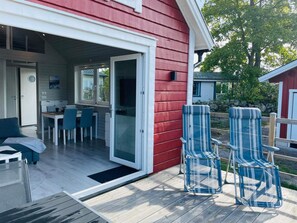 A red building with a covered outdoor area, a dining table with chairs, and two striped chairs on a deck.