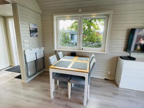 A room with a wooden table, two chairs, a window with a view of greenery, a television on a stand, and a white cabinet. - 4 Star Holiday Home in Solvesborg (Sölvesborg)