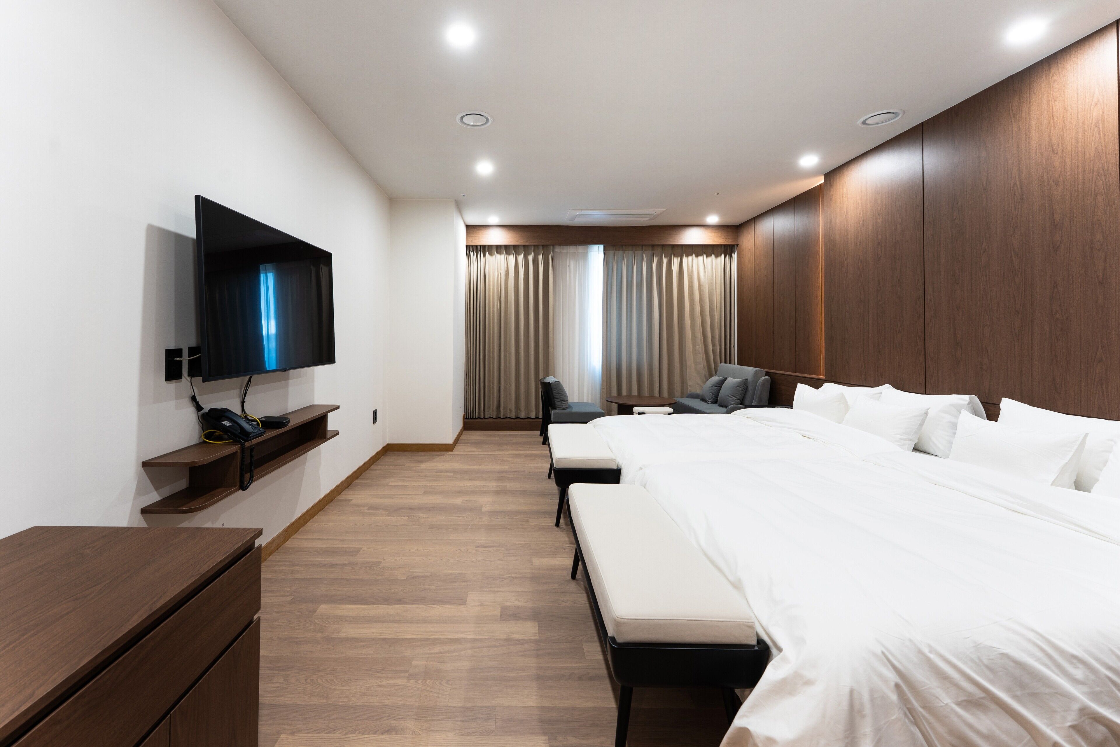 A modern hotel room with a large bed, a flat-screen TV mounted on the wall, a desk with a chair, and a window with curtains.