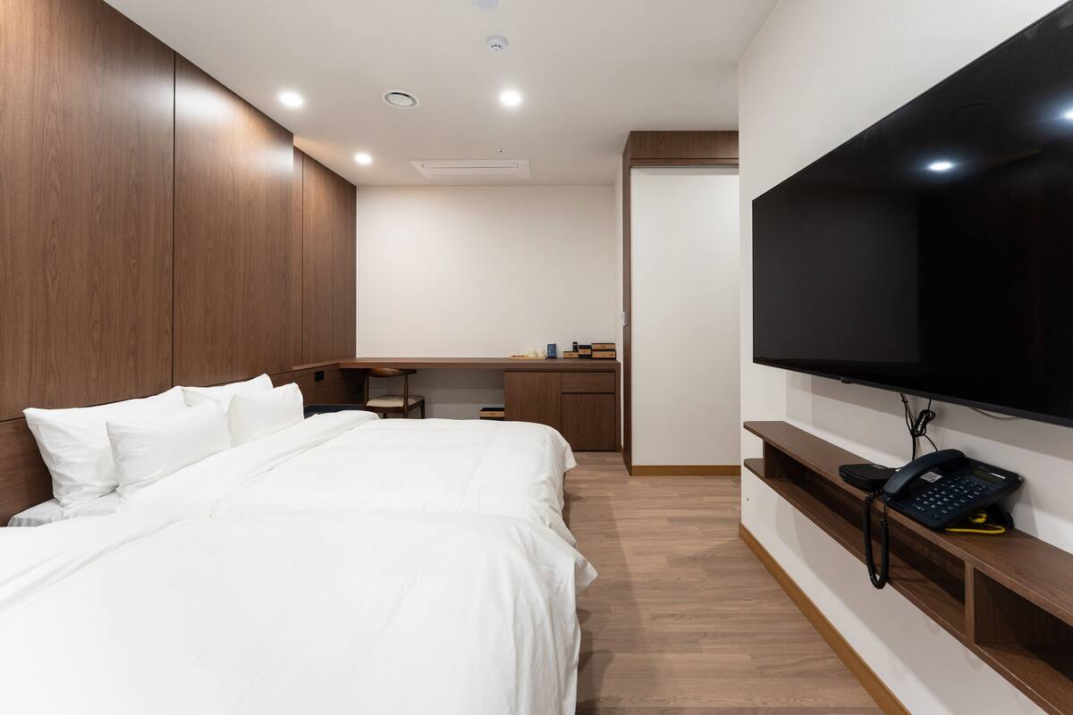 A hotel room with a large bed, a desk with a chair, a flat-screen TV mounted on the wall, and a telephone on a wooden shelf.