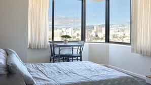 Ein Schlafzimmer mit einem großen Erkerfenster, einem Bett, einem kleinen Tisch mit Stuhl und einer Zimmerpflanze.