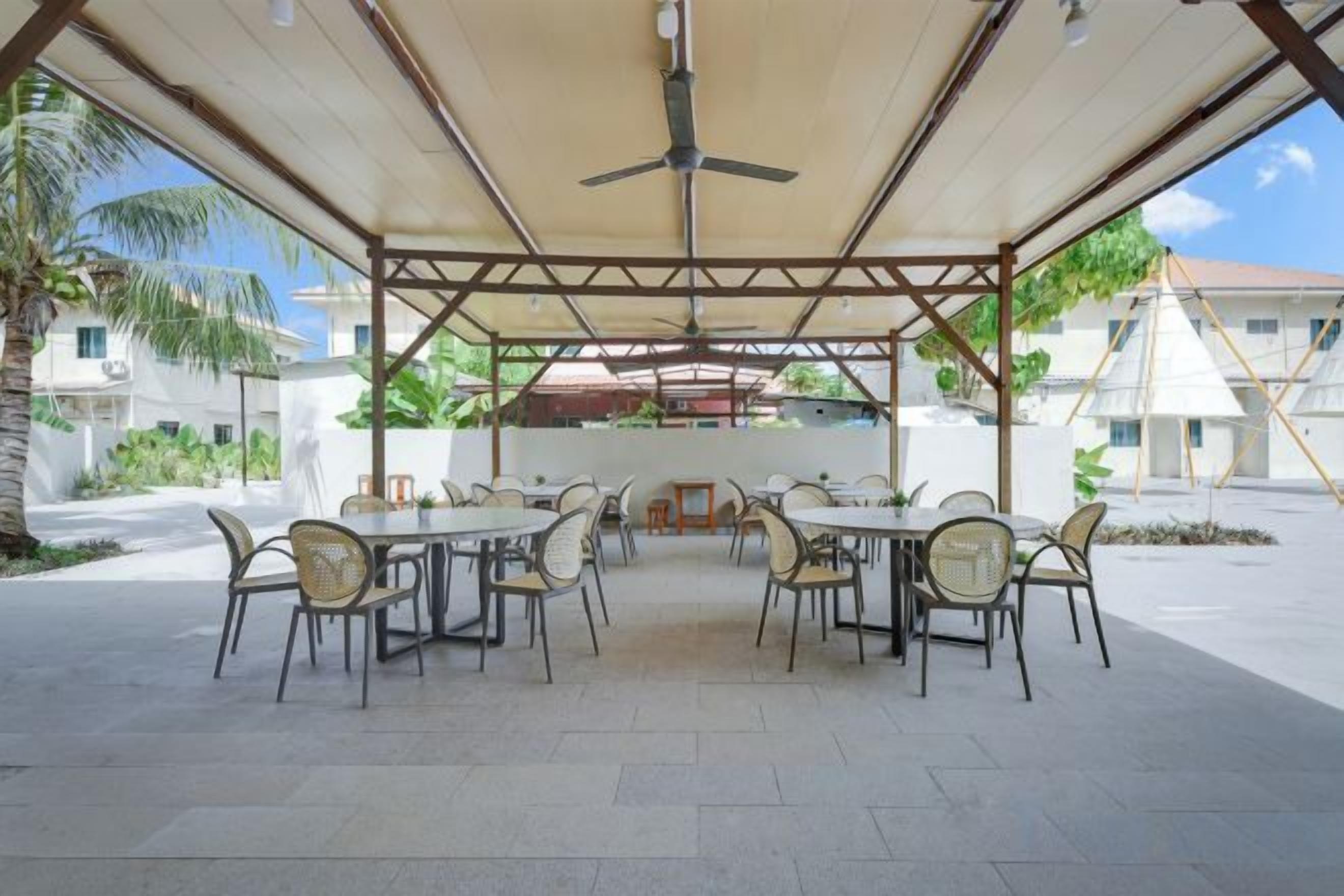 Une terrasse extérieure avec des tables et des chaises, un ventilateur de plafond et une vue sur les bâtiments et la verdure.