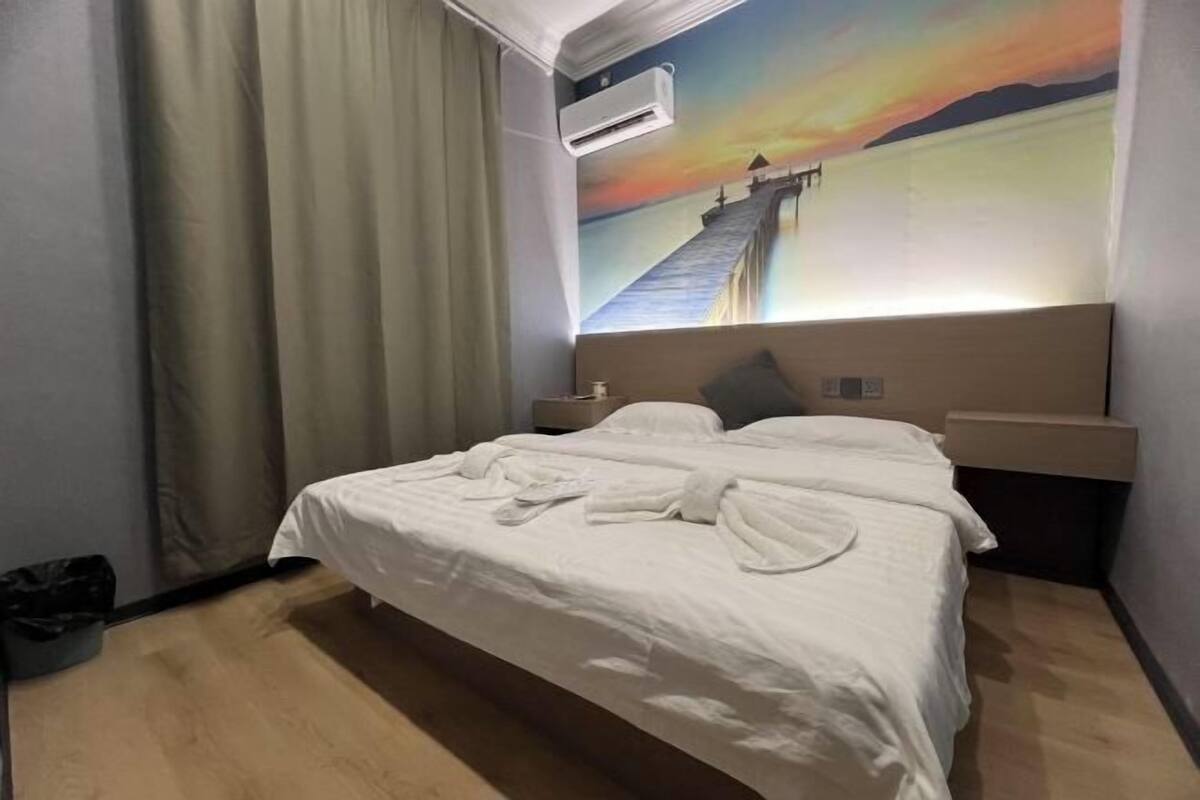 Une chambre d’hôtel avec un lit, une table de chevet, un climatiseur fixé au mur et un grand tableau représentant un coucher de soleil sur une jetée.