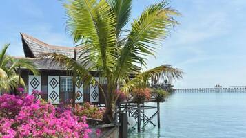A traditional overwater bungalow with a wooden deck, surrounded by water and palm trees.