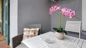 Un coin de pièce avec une table blanche, une chaise en osier, un coussin et un pot de fleurs contenant une plante aux fleurs roses.