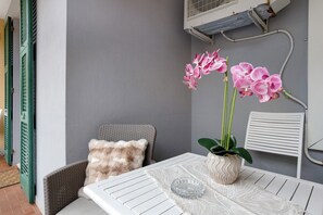 A corner of a room with a white table, a wicker chair, a cushion, and a potted plant with pink flowers.