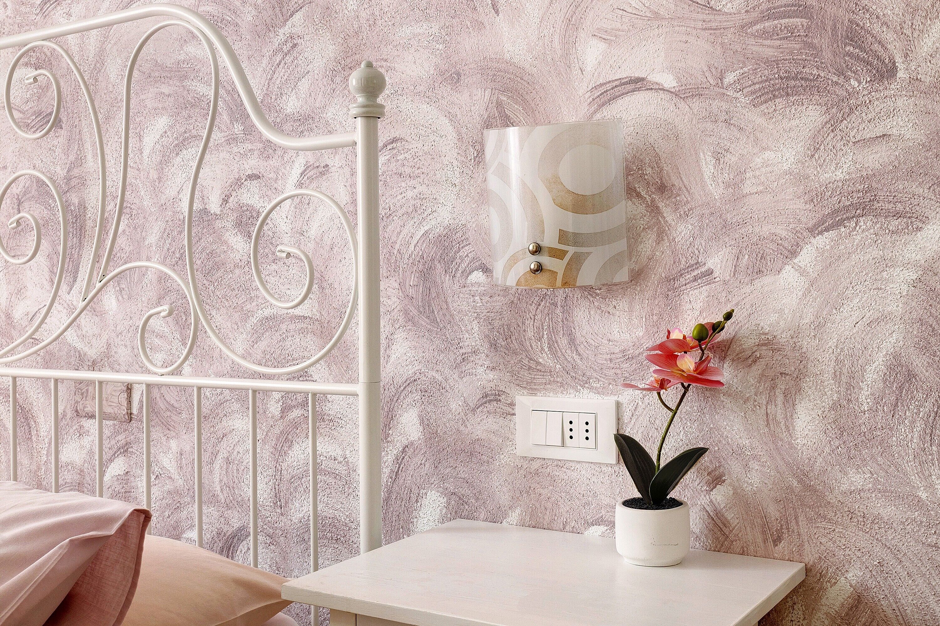 Una cama con cabecera de metal, una mesita de noche con planta en maceta, una lámpara de pared y un enchufe.