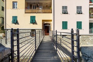 Un chemin de pierre mène à un bâtiment doté d’un balcon et de nombreuses fenêtres équipées de volets.