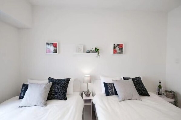 Habitación de hotel con dos camas, cada una con ropa de cama blanca y almohadas estampadas. Hay repisas de pared con objetos decorativos e imágenes enmarcadas.