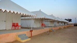 A row of white tents with a flat roof, each with a small porch and red chairs. - Jodhani Desert Camp & Resort Jaisalmer (Jaisalmer)