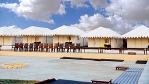 A row of white tents with wooden furniture and a flat surface in the foreground. - Jodhani Desert Camp & Resort Jaisalmer (Jaisalmer)