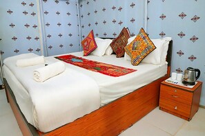 A neatly made bed with white linens, a red and green bedspread, and colorful pillows. There is a wooden nightstand with a kettle and cups beside the bed.