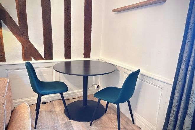 Une pièce d’angle avec une table ronde et deux chaises, des poutres en bois au mur et un rideau bleu.