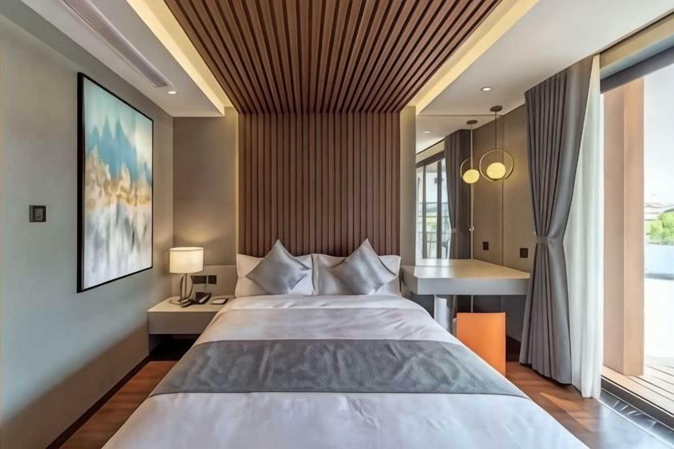 Una habitación de hotel moderna con una cama grande, una mesita de noche, una lámpara de pared y un amplio ventanal con cortinas.