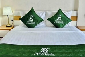 A hotel room with a neatly made bed, two green pillows with the hotel's logo, and a matching green throw blanket. - The Shilla Hotel Phu My Hung (Ho Chi Minh City)