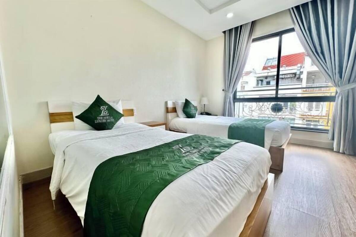 Una camera d'albergo con due letti, ognuno con una coperta verde e cuscini coordinati, un'ampia finestra con tende e vista sugli edifici circostanti.