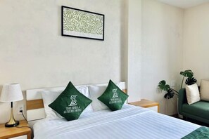 A hotel room with a bed, two pillows with a logo, a nightstand with a lamp, a green sofa, and a plant.