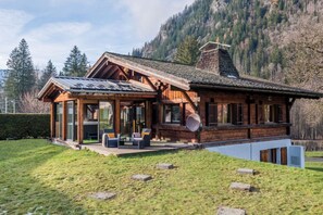 A wooden cabin with a glass extension, a deck, and a mountainous backdrop.