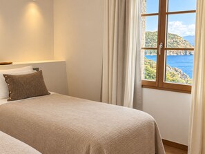 A bedroom with a bed, a nightstand, a lamp, and a window with a view of a coastal landscape.