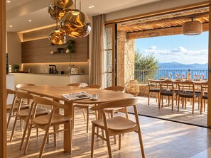 A modern kitchen with wooden cabinets and a stone wall, a dining area with a wooden table and chairs, and a balcony with a view of the sea. - Villa Lunassina (Cargèse)