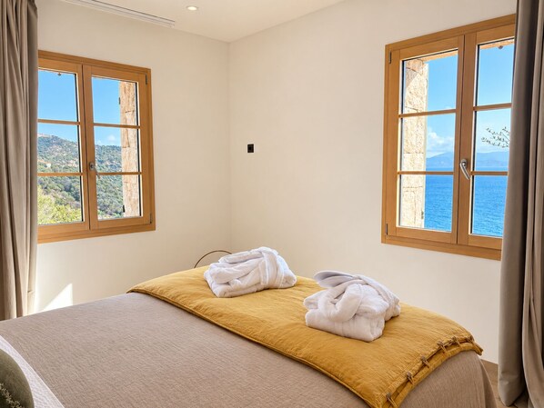 A hotel room with a bed, two windows with wooden frames, and a view of the sea.