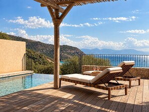 A wooden deck with lounge chairs and a pool, overlooking a mountainous landscape and the sea.