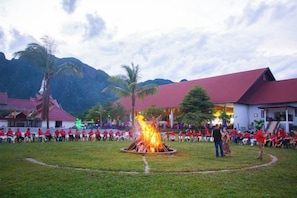 A group of people gathered around a fire pit in an outdoor setting with buildings in the background. - Hometown of Dragon Hotel (Vang Vieng)