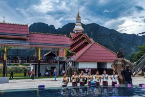 A traditional building with a red roof and a golden spire, a pool area with people, and a large elephant statue. - Hometown of Dragon Hotel (Vang Vieng)