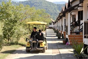A yellow vehicle with two passengers on a paved path beside a building with a staircase leading to an upper level.