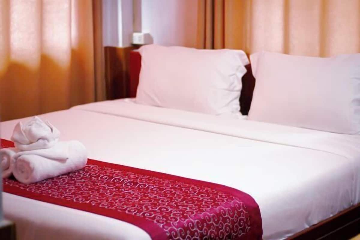 Cama bem arrumada com lençóis brancos e colcha vermelha estampada, toalha branca dobrada e cabeceira de madeira.