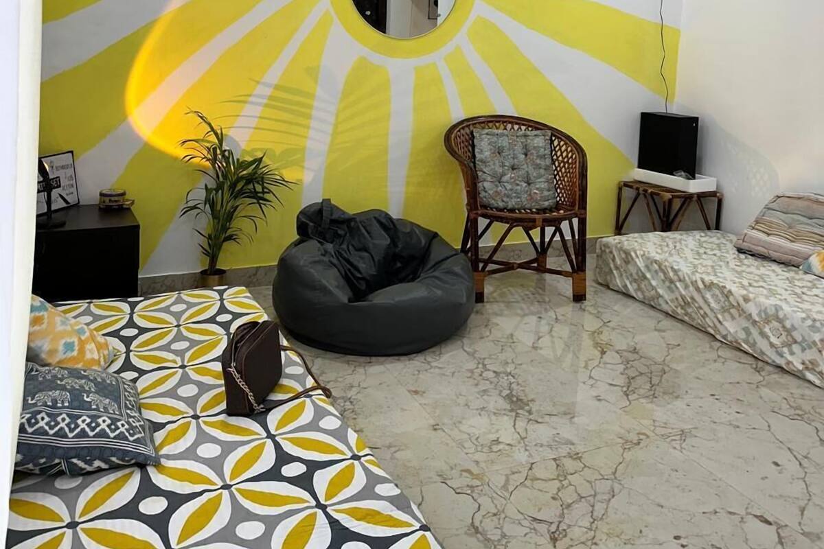 Una camera da letto con un murale a forma di raggio di sole, un letto con lenzuola a fantasia, un pouf, una sedia e una TV su un mobile.