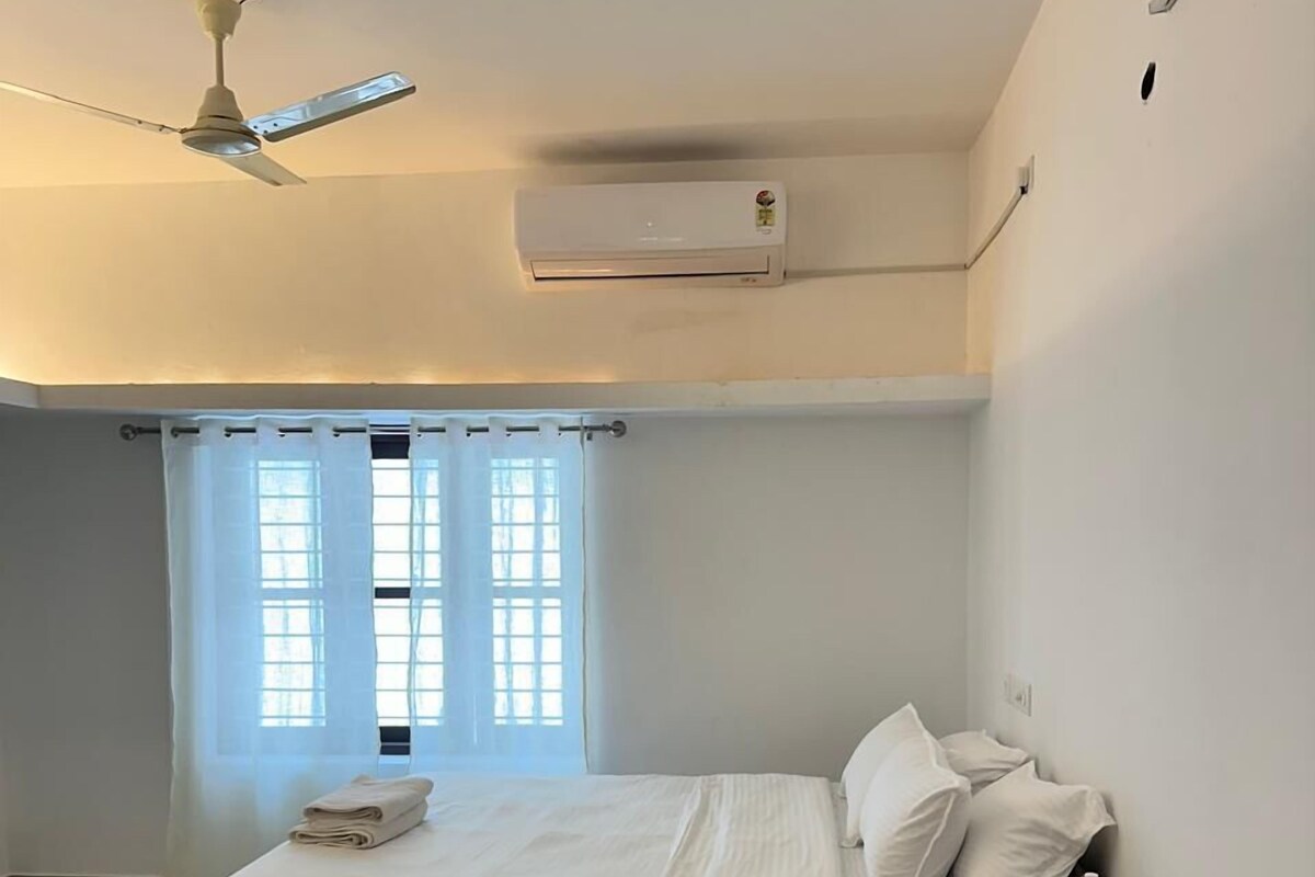 Et soveværelse med seng, et natbord, en lampe, et vindue med gardiner, en loftsvifte og en aircondition.