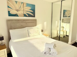 A neatly made bed with white linens and two rolled towels, a bedside table with a lamp, and a large mirror reflecting the room.
