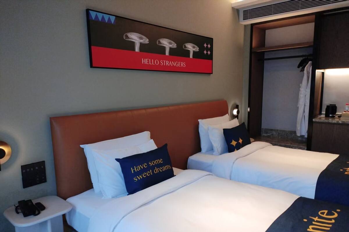 Ein Hotelzimmer mit zwei Betten, einem Kopfteil, einem Nachttisch mit Lampe, einem Wandbild und einem Kleiderschrank mit Kleidung.