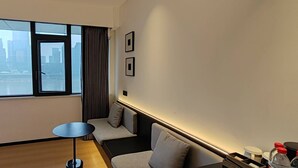 A hotel room with a large window offering a city view, a small round table, a sofa, and a wall-mounted shelf with bottles. - Chongqing Justom Family Apartment Hotel (Chongqing)