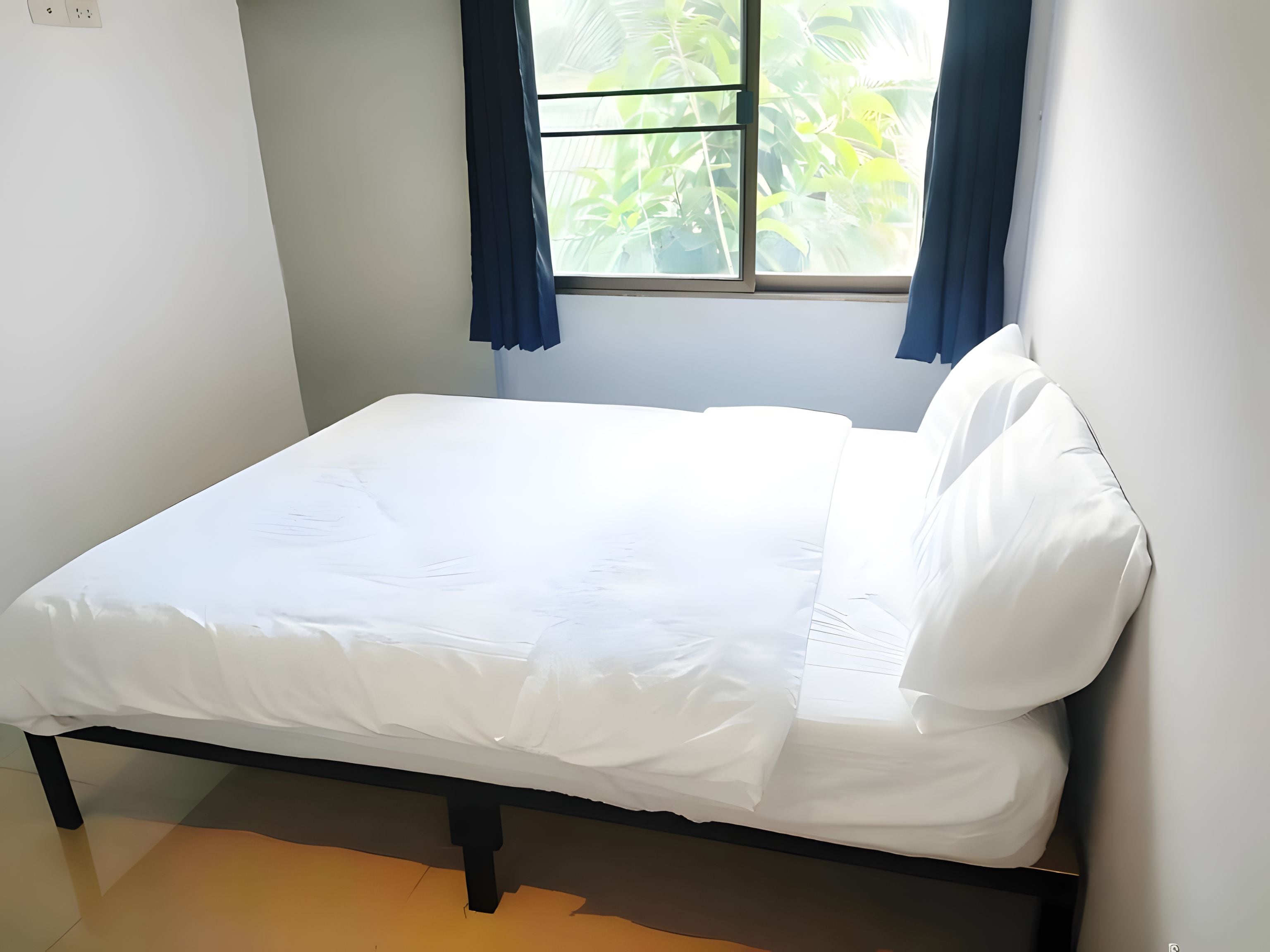 A single bed with a white mattress and pillows, positioned against a wall with a window above.