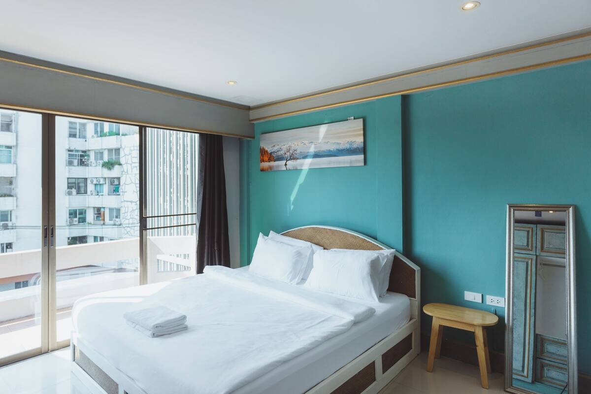 A modern hotel room with a large bed, a small bedside table, a painting on the wall, and a view of a cityscape through large windows.