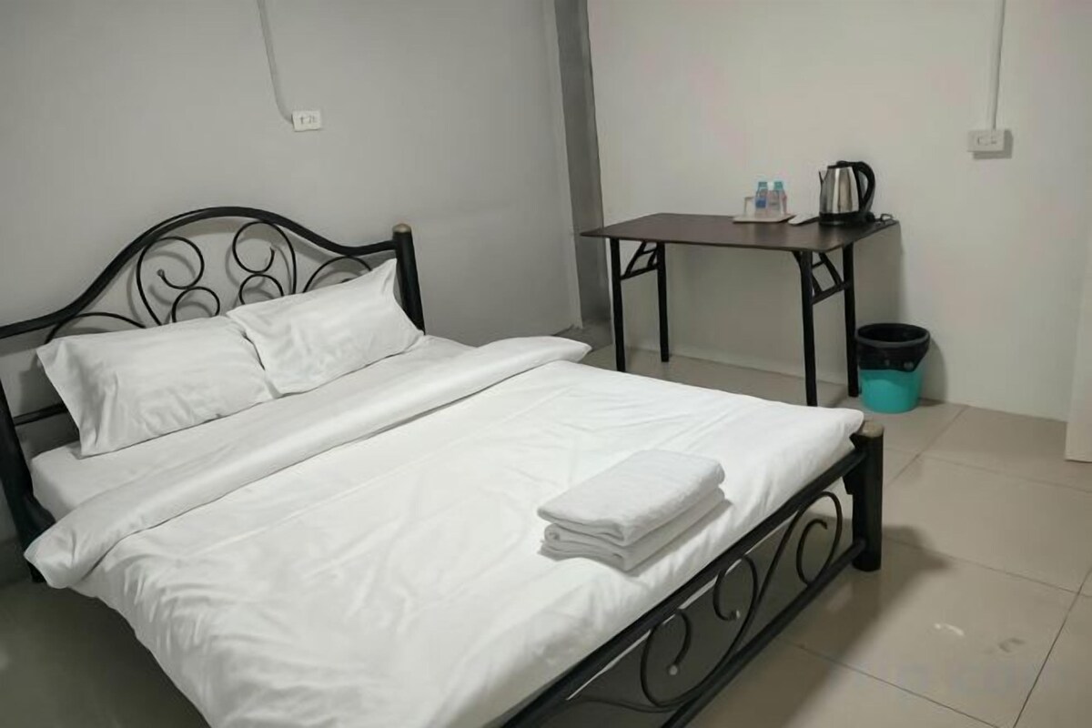 Een keurig opgemaakt bed met witte lakens, een metalen hoofdbord en een klein tafeltje met een waterkoker en kopjes ernaast.