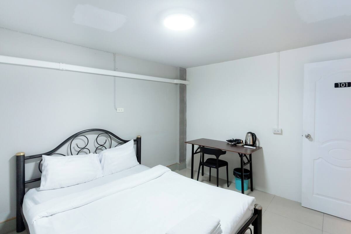 A room with a bed, a desk, a chair, a kettle, and a door with a number.