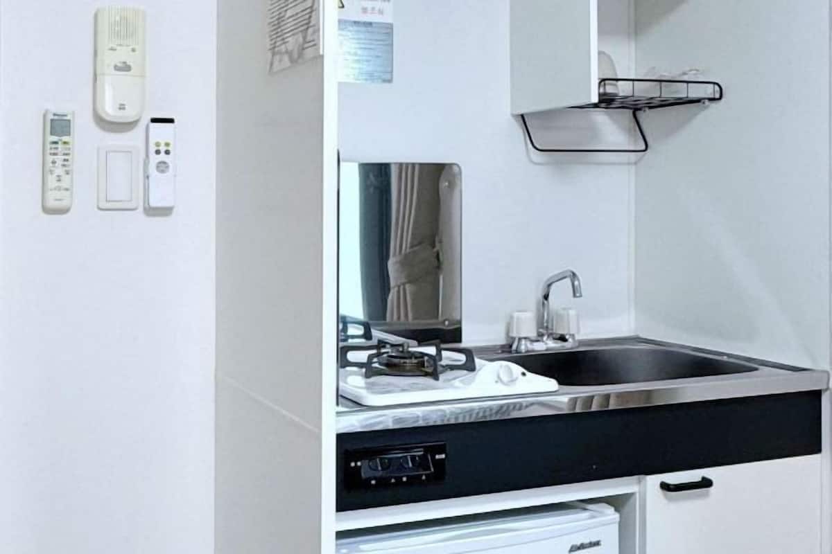 A compact kitchen with a sink, stove, and a small countertop. There are three remote controls on the wall, a black towel, and a white cabinet below the countertop.