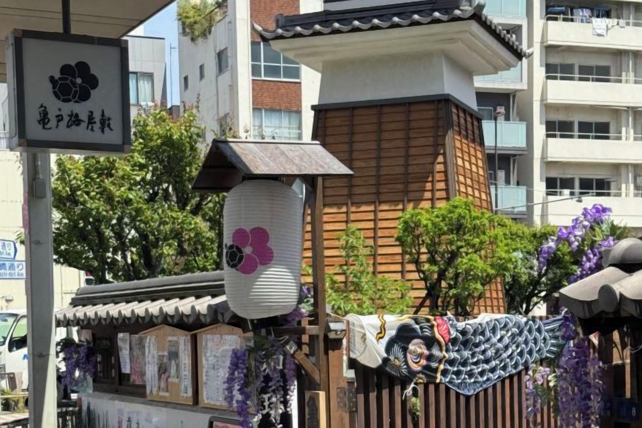 Een traditioneel Japans gebouw met een betegeld dak, een bord met Japanse karakters en een klein bouwwerk met bloemdecoratie in paars.
