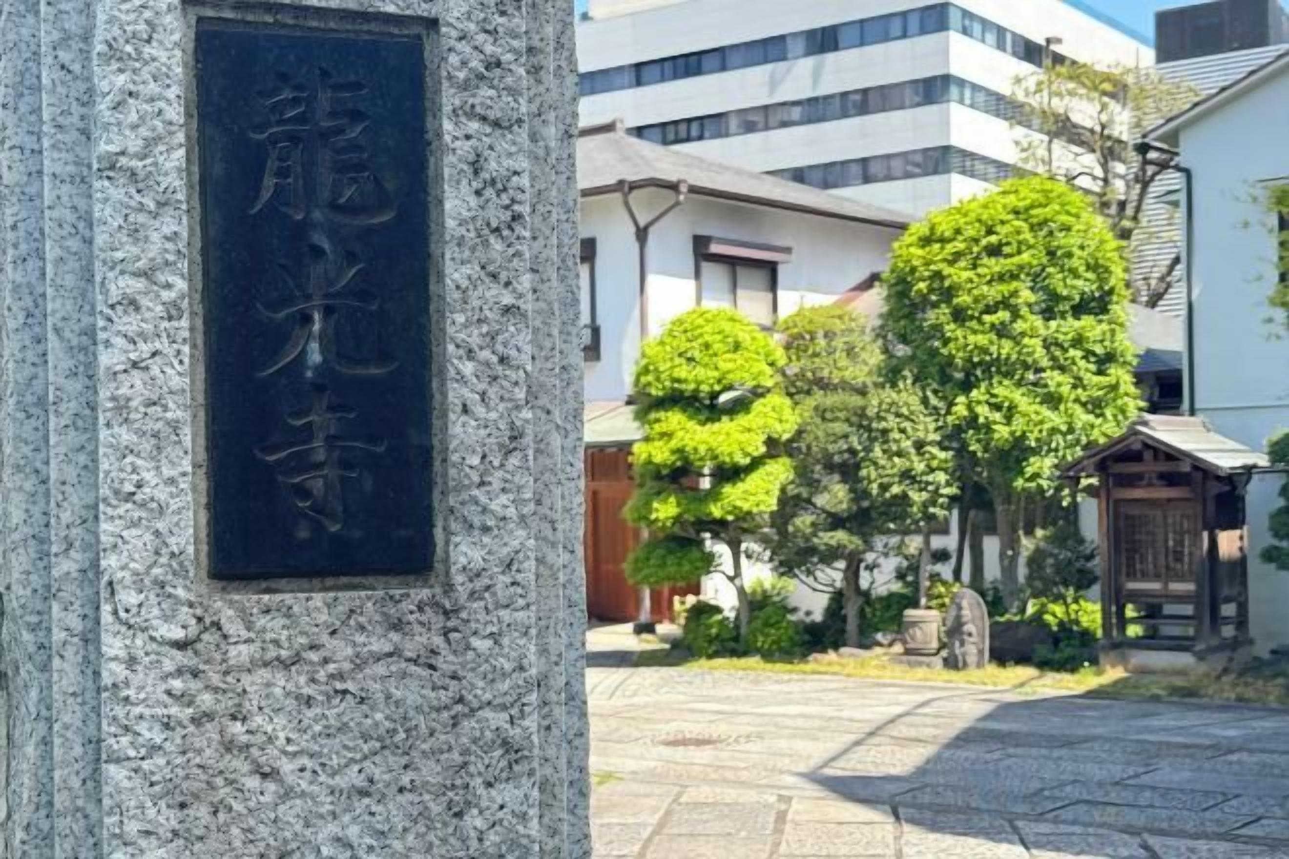 Monument de pedra amb caràcters japonesos, un pati pavimentat i edificis tradicionals al fons.