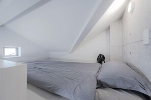 Een slaapkamer met een schuine kap, een bed met grijze beddengoed en een klein raam.